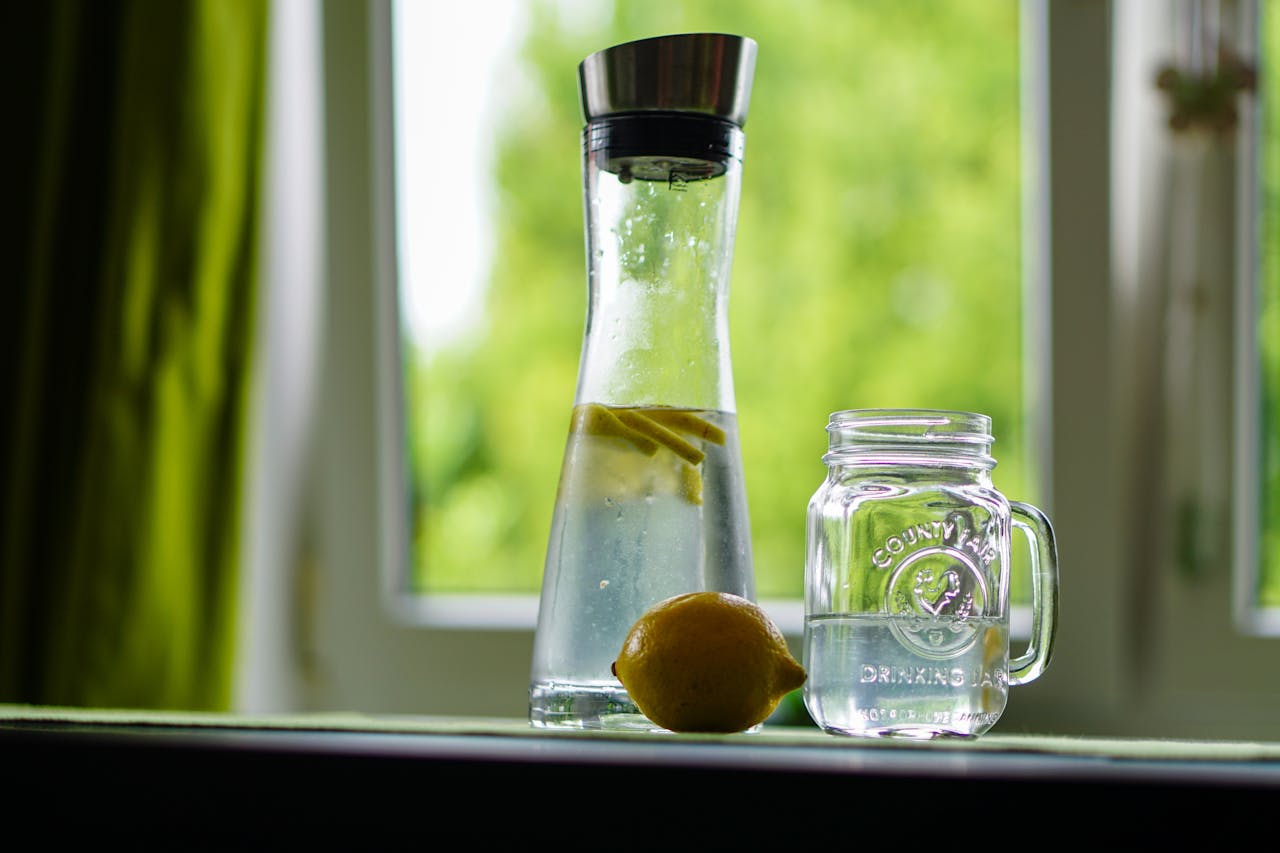 Lemon-infused water in a glass carafe and mason jar on a windowsill, evoking freshness.