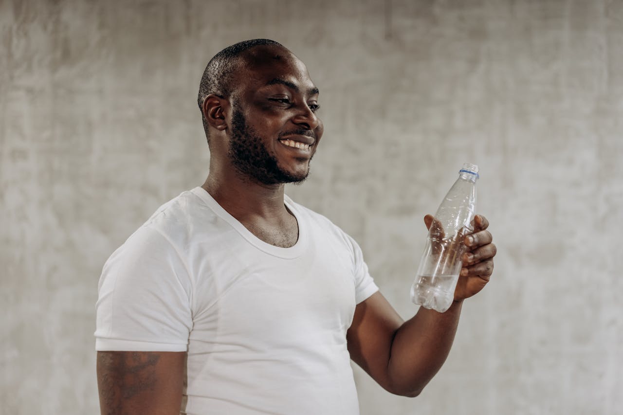 A happy man smiles while holding a bottle of water indoors, embracing healthy living.